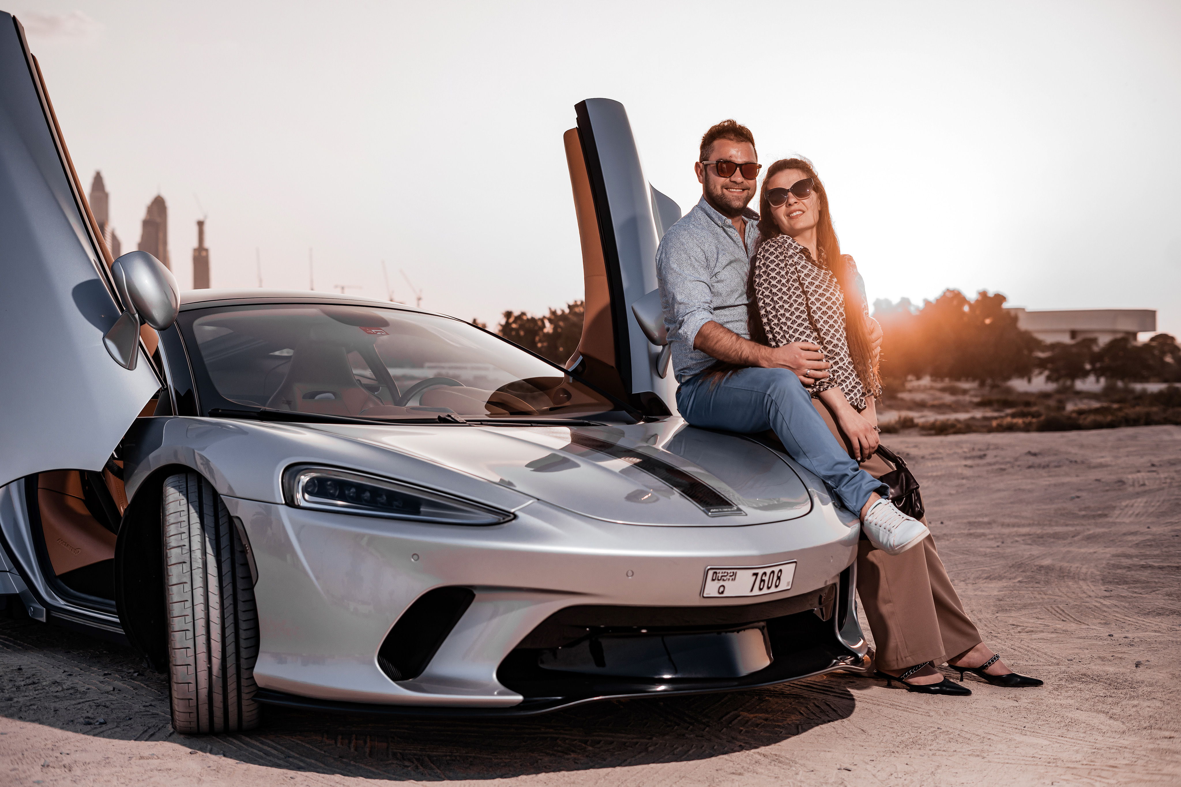 A man and woman pose next to a luxury silver sports car during a photoshoot in Dubai.